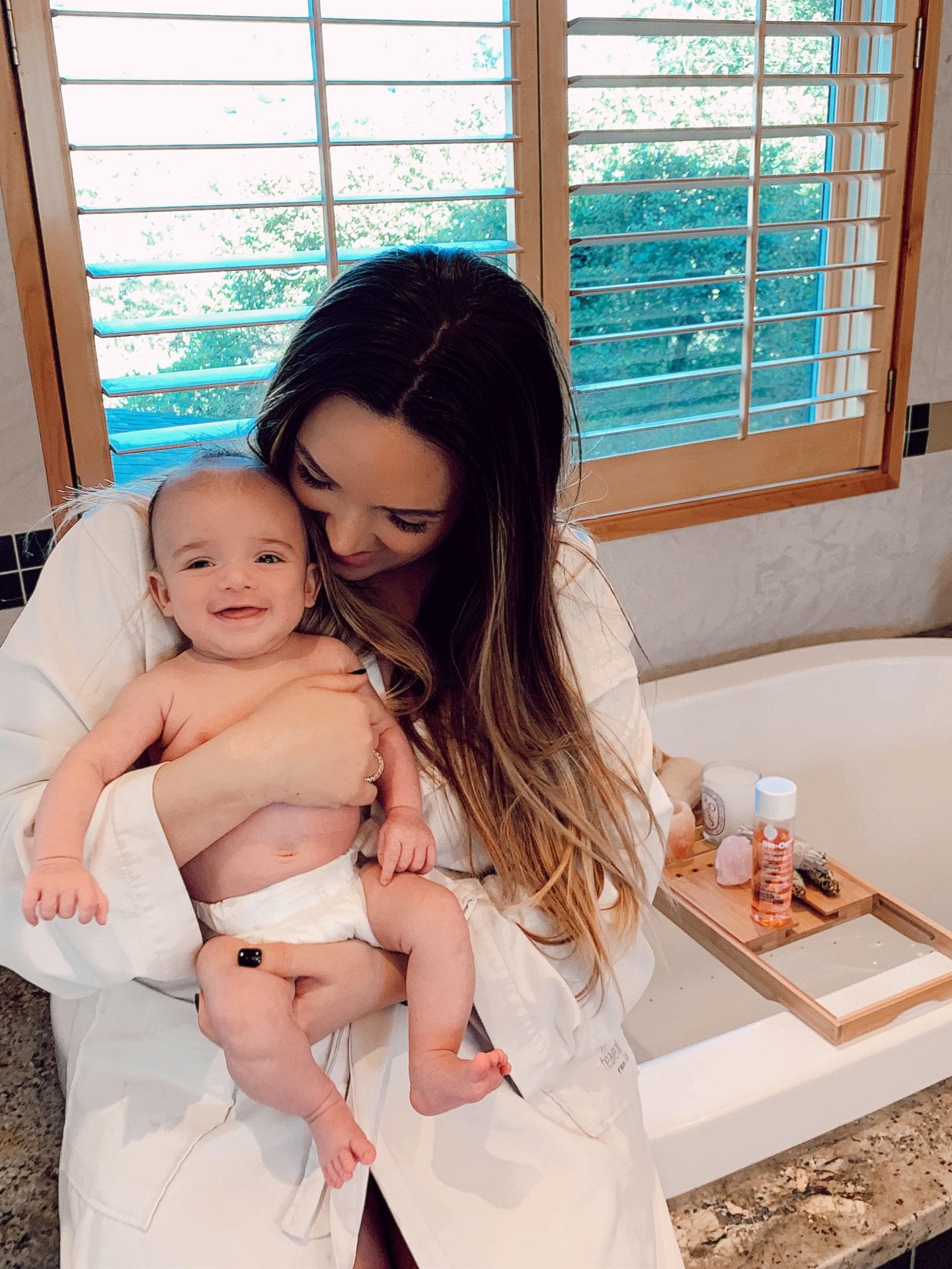 Bio Oil for Stretch Marks by popular San Francisco lifestyle blog, Just Add Glam: image of a mom wearing a white bathrobe and sitting on the edge of her tub while holding her baby.  