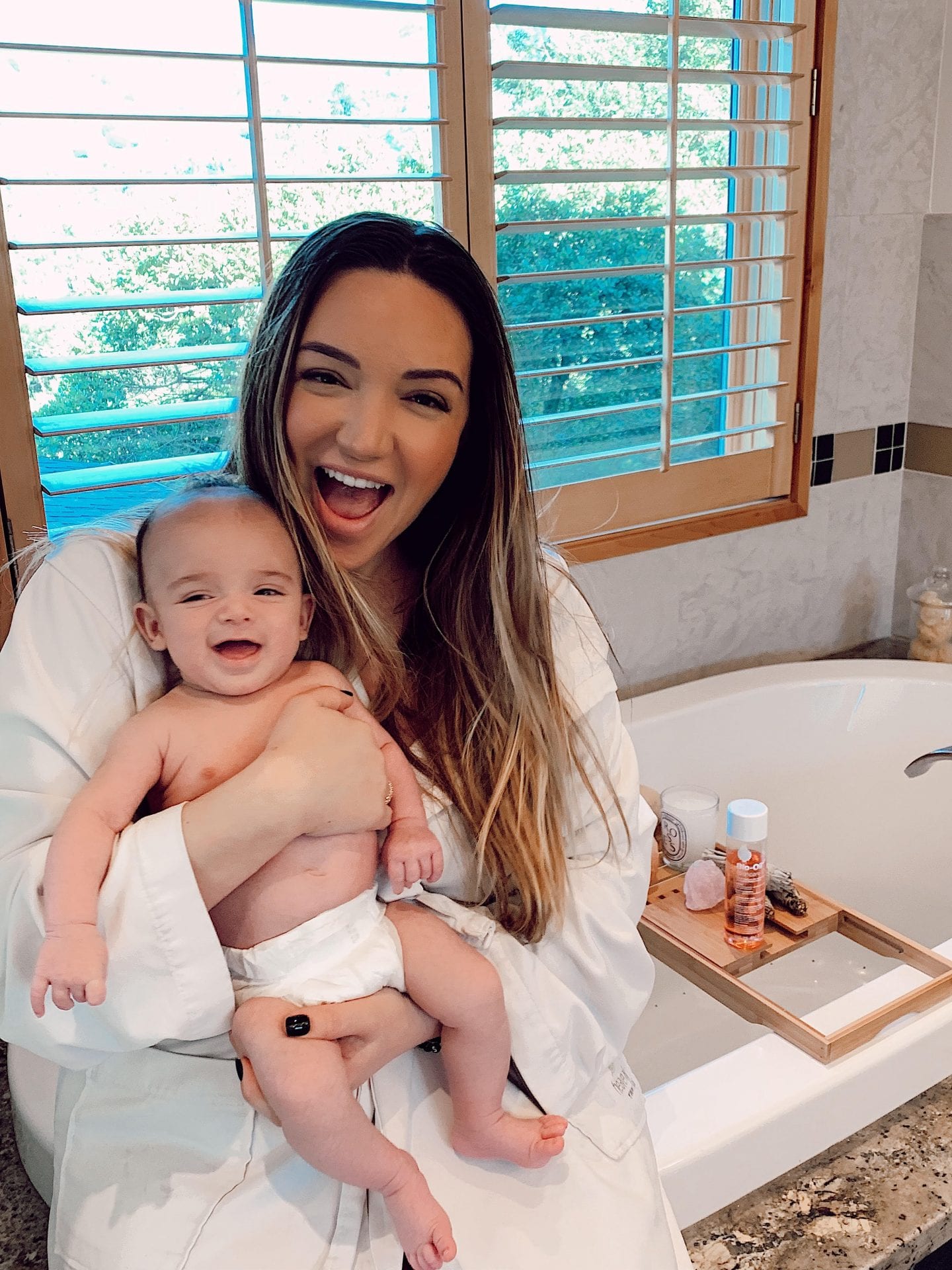Bio Oil for Stretch Marks by popular San Francisco lifestyle blog, Just Add Glam: image of a mom wearing a white bathrobe and sitting on the edge of her tub while holding her baby.  